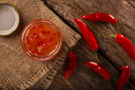 A jar of red pepper jelly on a wooden table with red peppers